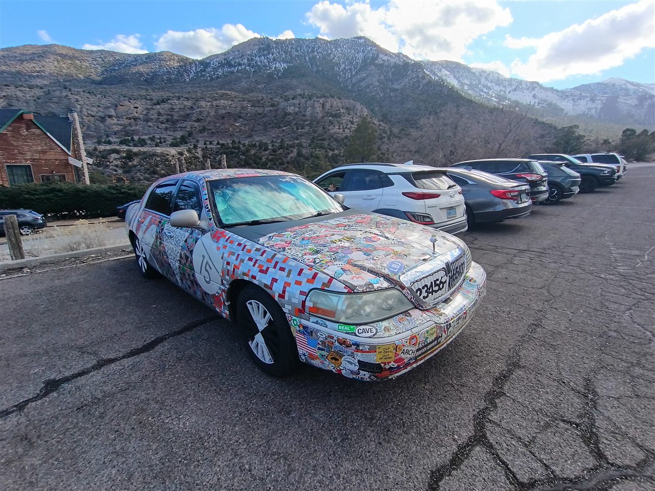 A car completely covered in colorful stickers parked in a mountain lot with snow-capped peaks behind it