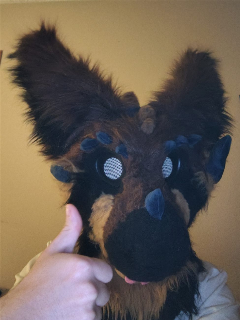 A brown and tan fursuit head with blue felt spikes and mesh eyes, thumbs up