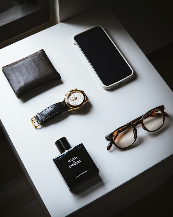 A bottle of Bleu de Chanel on a table with a watch, wallet, and glasses